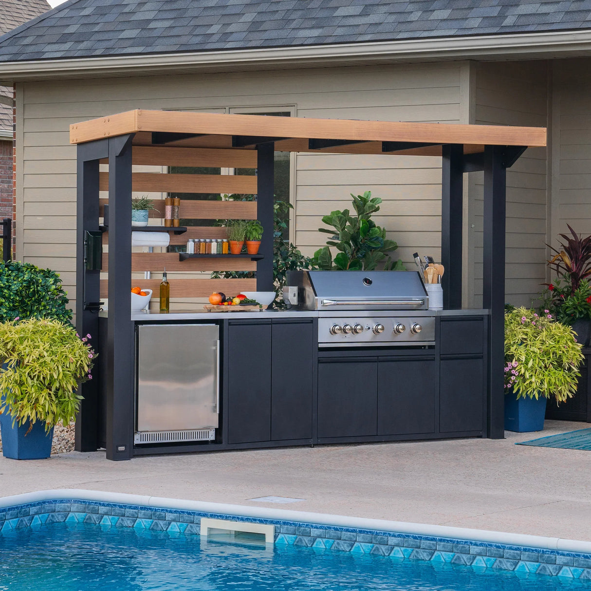 Modern outdoor kitchen with pergola, grill, stainless fridge, and plants by a pool