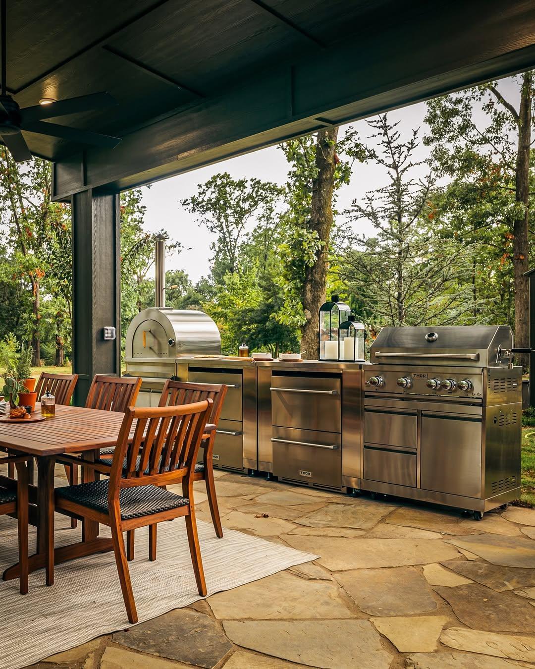 Outdoor kitchen with stainless steel grill, pizza oven, and wooden dining table set on stone patio