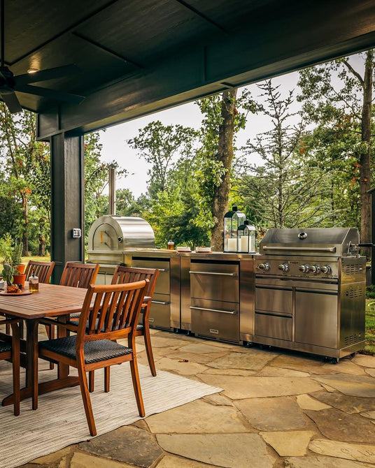 Outdoor kitchen with stainless steel grill, pizza oven, and wooden dining table set on stone patio