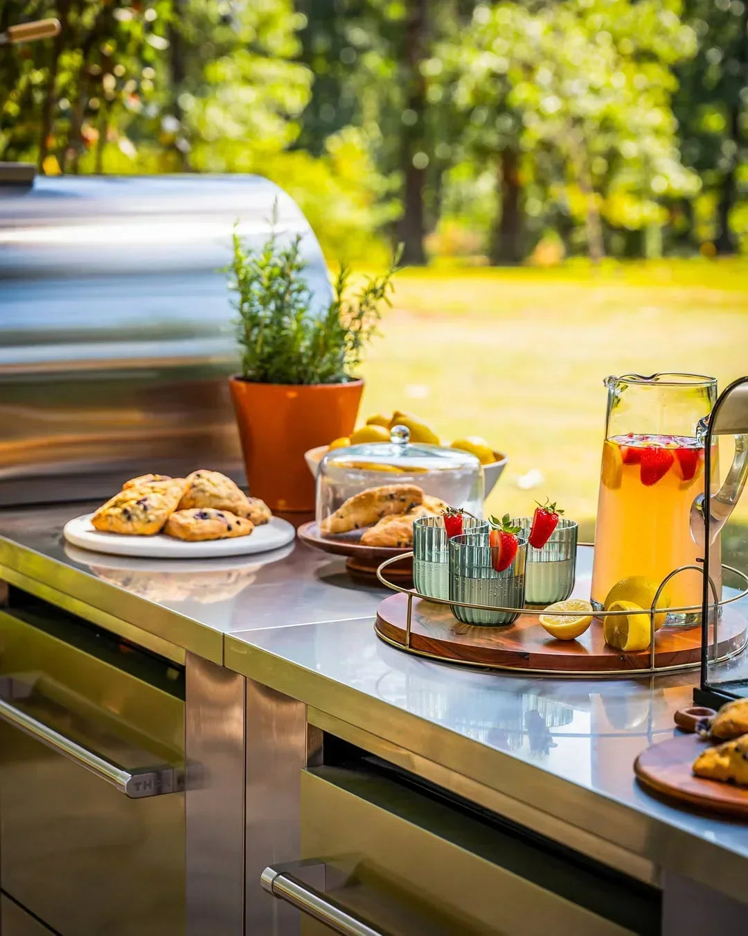 Outdoor kitchen counter with pastries, strawberry lemonade, and potted herbs in sunlight