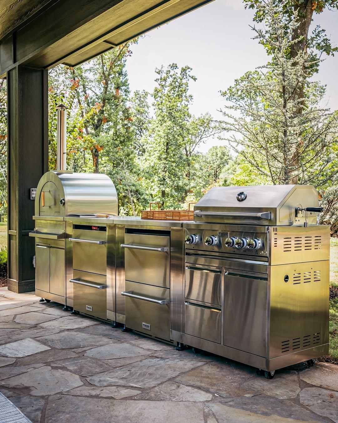 Stainless steel outdoor kitchen with grill, pizza oven, and storage units on stone patio