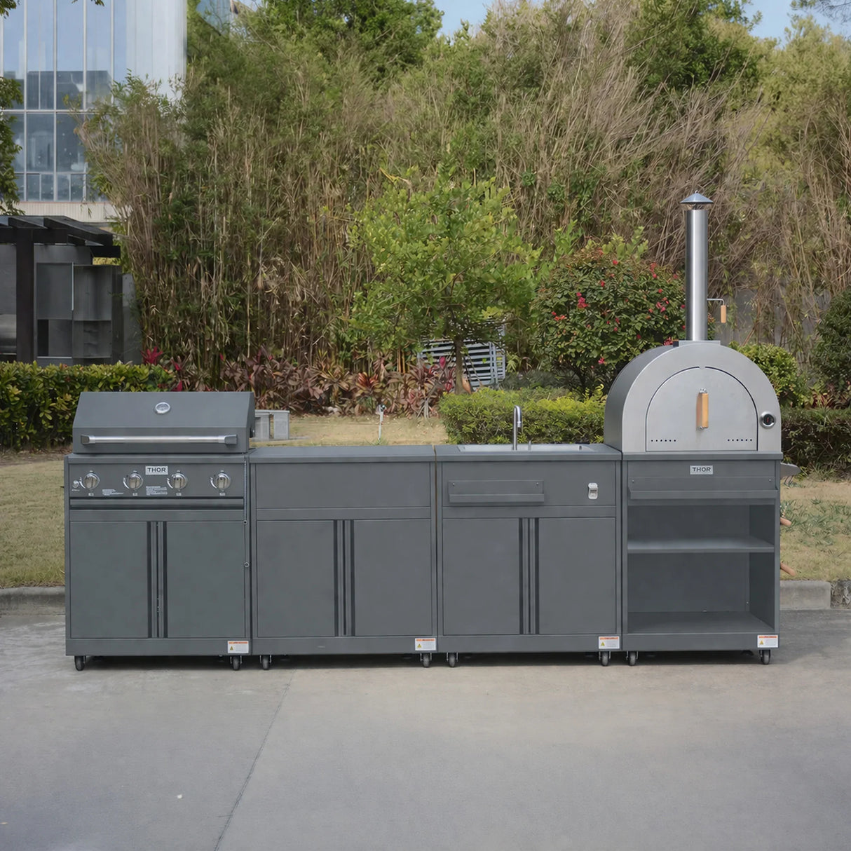 Modern outdoor kitchen set with grill, pizza oven, sink and storage, in a garden setting.