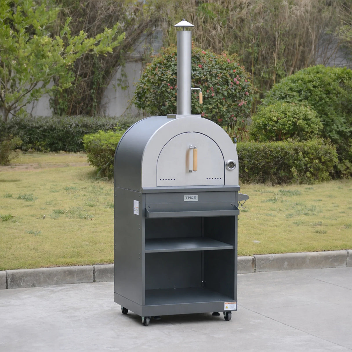 Outdoor stainless steel pizza oven with chimney and shelves on wheels in a garden setting