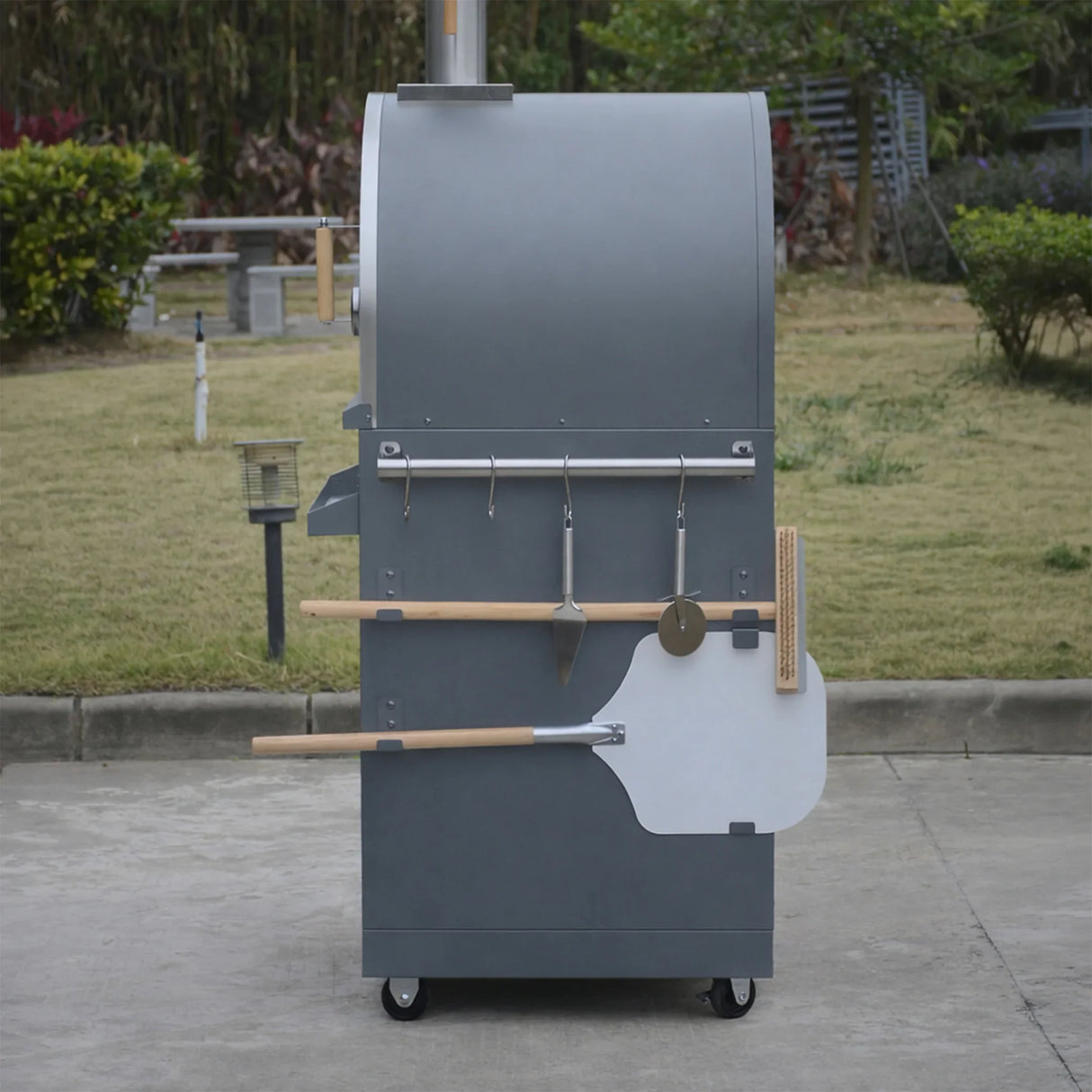 Outdoor wood-fired pizza oven with tools on wheels in a garden setting