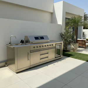 Modern stainless steel outdoor kitchen with grill, sink, fridge on patio by white house